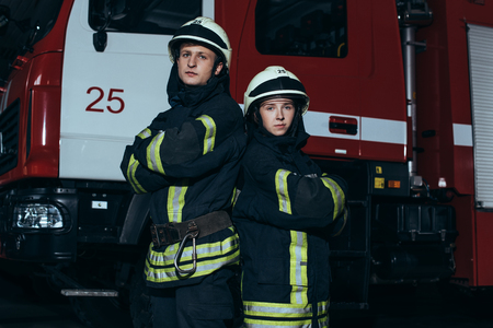 Portrait of firefighters in fireproof uniform and helmets with arms crossed standing near truck at fire stationの写真素材