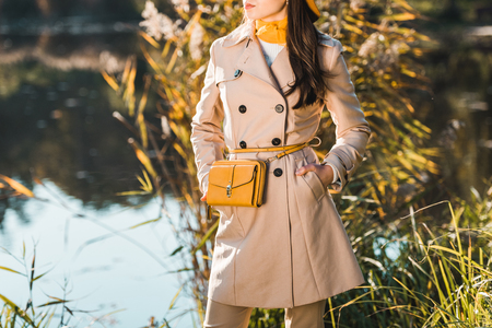 Partial view of stylish woman in trench coat posing with yellow bag near pond in parkの写真素材