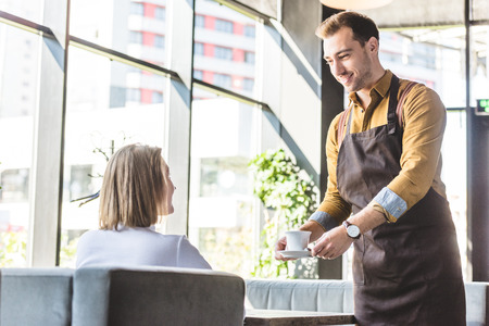 Attractive young waiter serving cup of coffee for female client at cafeの写真素材