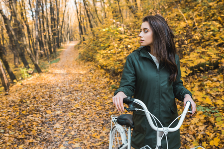 Selective focus of beautiful young woman carrying bicycle in yellow autumnal forestの写真素材