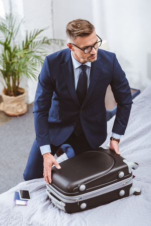 High angle view of businessman in suit and eyeglasses packing suitcase and looking away at homeの写真素材
