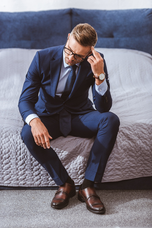 High angle view of thoughtful businessman in suit and eyeglasses sitting on bed at homeの写真素材