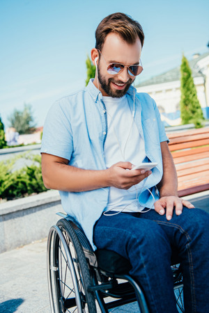 Smiling handsome man in wheelchair using smartphone and listening to music on streetの写真素材
