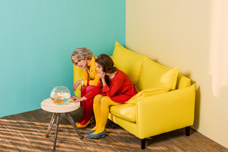 Retro styled women sitting on yellow sofa with aquarium fish on coffee table at colorful apartment, doll house conceptの写真素材
