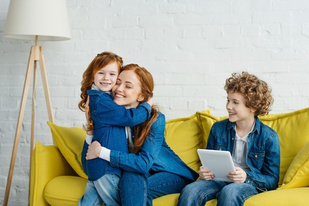 Son holding tablet while sitting on sofa with hugging mother and daughterの写真素材