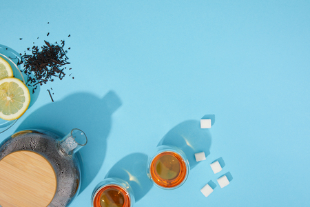 Top view of fresh tea in cups and teapot, sugar cubes, dry herbal tea and sliced lemon on blue backgroundの写真素材