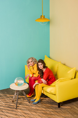 Retro styled women sitting on yellow sofa with aquarium fish on coffee table at colorful apartment, doll house conceptの写真素材