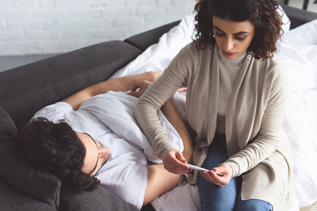 Girlfriend checking temperature of sick boyfriend with thermometer at homeの写真素材