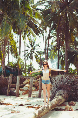 Young woman in sunglasses walking on fallen palm tree on beachの写真素材