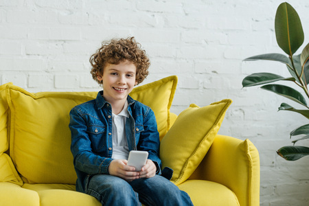 Smiling little boy using phone sitting on sofaの写真素材