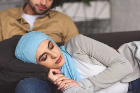 Muslim girlfriend in hijab sleeping on boyfriends legs at home