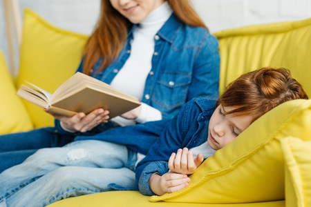 Mother reading book to sleeping daughter on yellow sofaの写真素材