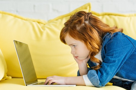 Cute redhead child using laptop on sofa