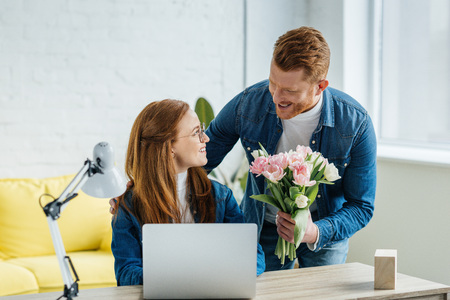 Man surprising young woman working by laptop with a bouquet of tulipsの写真素材