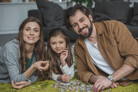 Smiling parents and daughter lying on carpet with puzzle pieces and looking at cameraの写真素材