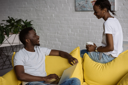 Smiling young man using laptop and looking at beautiful girlfriend holding cup of tea at homeの写真素材