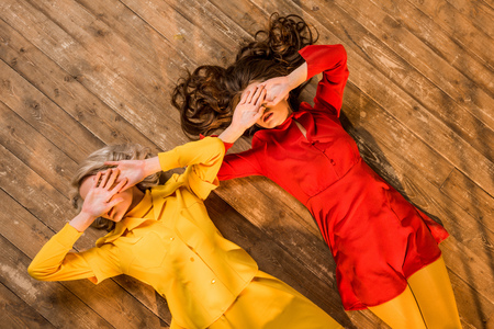 Top view of retro styled girls in colorful dresses lying on floor and covering eyes with hands at...