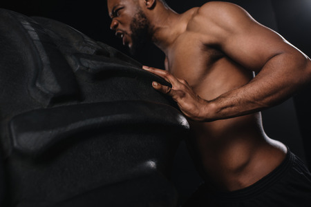 Low angle view of muscular young African American sportsman lifting tire on black backgroundの写真素材