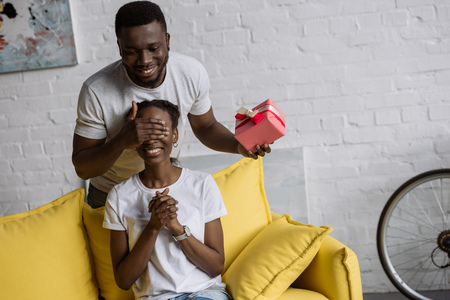 Smiling young African American man presenting gift box to happy girlfriend at homeの写真素材