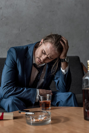Drunk depressed businessman sitting on couch with glass of whiskey on tableの写真素材
