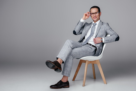 Smiling adult businessman in stylish suit sitting on chair on grey backgroundの写真素材
