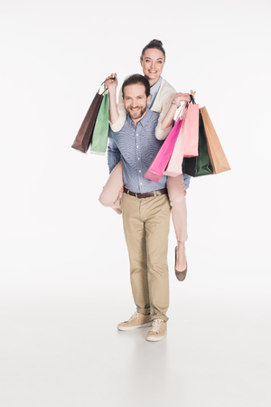 Happy woman with shopping bags and husband piggybacking together isolated on white backgroundの写真素材