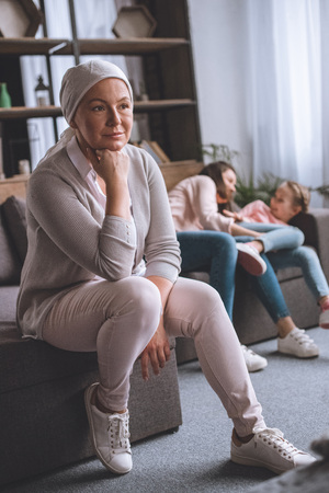 Pensive sick mature woman in kerchief looking away while family members having fun behindの写真素材