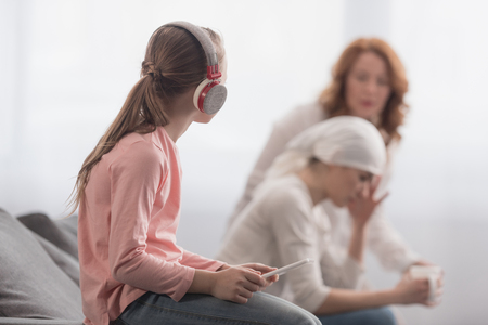 Child in headphones using digital tablet and looking at sick mother and grandmother talking behindの写真素材