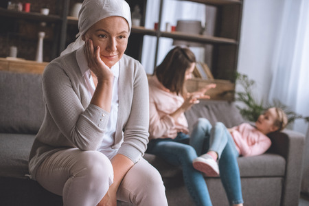 smiling sick mature woman in kerchief looking away while family members having fun behindの写真素材