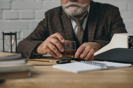 Cropped shot of senior bearded writer with glass of whiskey at workplaceの写真素材
