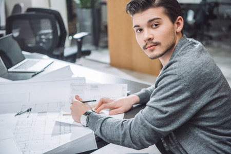 Handsome young architect drawing building plans at office and looking at cameraの写真素材