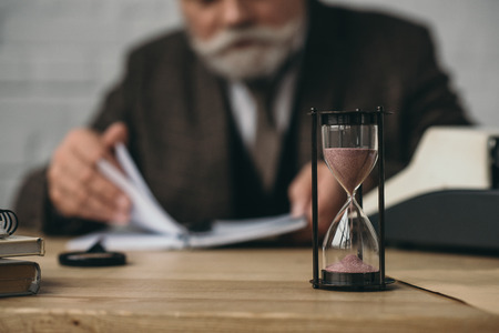 Senior writer working with hourglass standing on foreground