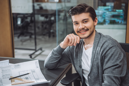Handsome young architect sitting at workplace with building plans at office