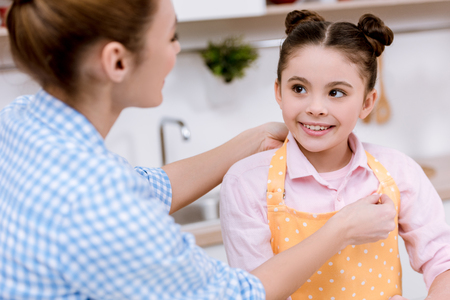 Mother dressing her daughter in apron at kitchenの写真素材