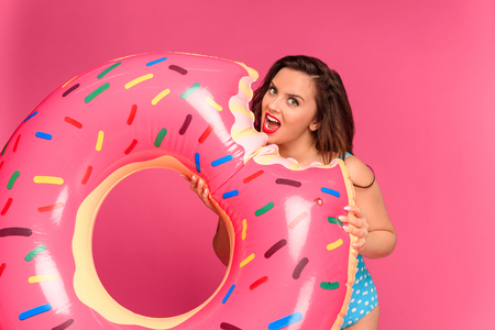 Young brunette woman in swimwear holding inflatable ring and looking at camera isolated on pink backgroundの写真素材