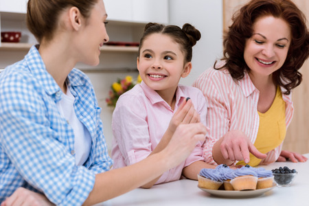 Three generations of women decorating cupcakes with blueberries together at kitchenの写真素材