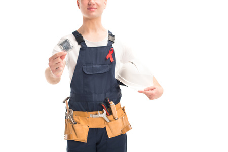 cropped image of worker in uniform and red ribbon holding helmet and condom isolated on white, world...