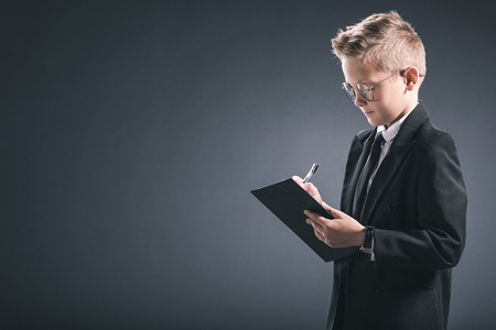 preteen boy in businessman suit and eyeglasses making notes on notepad on grey backgroundの写真素材