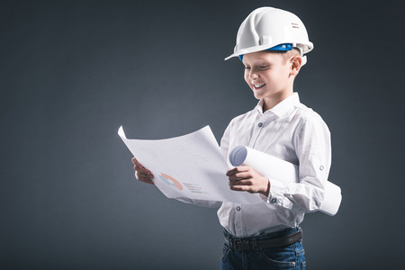 portrait of smiling boy in architect helmet looking at blueprints in hands on dark backgroundの写真素材