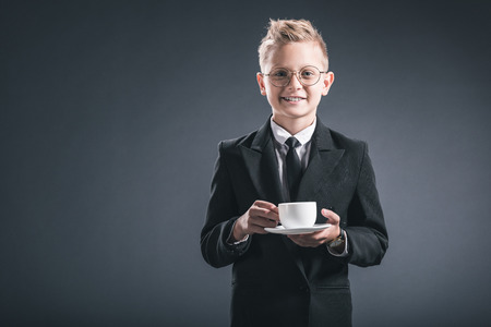 portrait of smiling boy in businessman suit and eyeglasses holding cup of coffee on dark backdropの写真素材