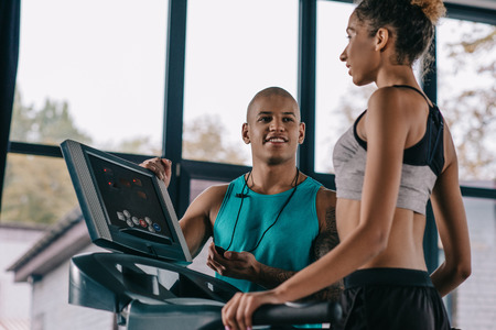 young male trainer with timer looking at sportswoman running on treadmill at gymの写真素材