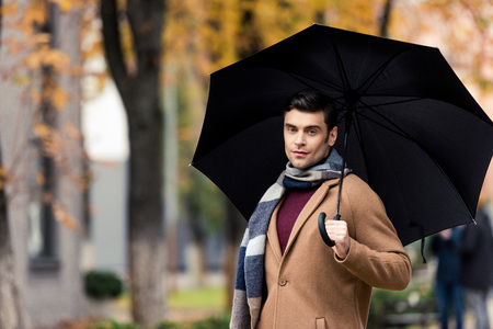 attractive stylish man in coat and scarf with umbrella walking by autumnal streetの写真素材