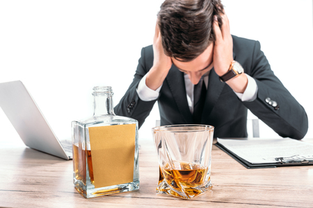 exhausted businessman touching head and leaning on table, whiskey on foreground isolated on whiteの写真素材