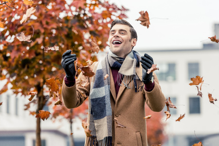handsome happy man in coat and scarf throwing up fallen leaves on streetの写真素材