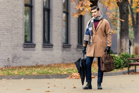 handsome man in coat with briefcase and umbrella walking by autumnal streetの写真素材