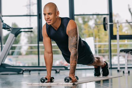 handsome african american sportsman with tattooed hand doing push ups with dumbbells on fitness mat...