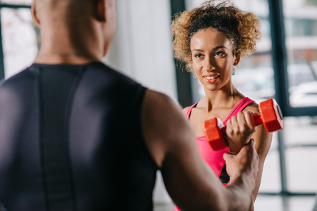 partial view of african american male trainer helping sportswoman to exercising with dumbbell at gym