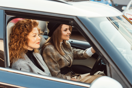 side view of happy stylish woman in black hat driving car while her ginger female friend sitting...