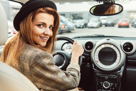 Adult cheerful woman in black hat and checkered jacket sitting at steering wheel in car