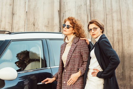 Selective focus of stylish adult women in coats and sunglasses posing near car at city streetの写真素材
